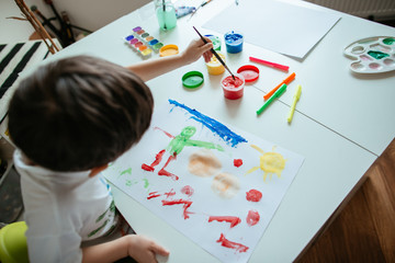 Top view of left handed 5 year old boy reaching for red paint with paintbrush.