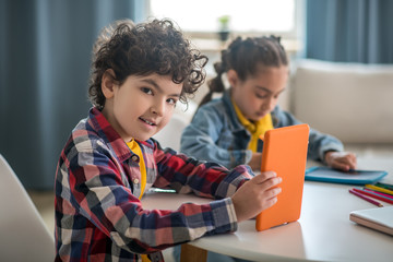 Boy and girl sitting at round table, holding tablets, busy with writing, boy looking into the camera