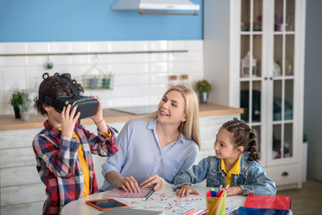 Curly boy and dark-haired girl sitting at round table with blonde female, boy wearing vr glasses