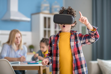 Curly boy wearing vr glasses, looking excited, dark-haired girl and blonde woman sitting at round table, busy with tasks