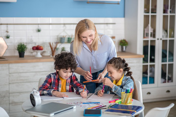 Curly boy and dark-haired girl sitting at table, doing their assignments, blonde female standing above them, helping