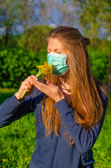 A young blond woman in a medical mask sniffs flowers in a Park. Spring, Allergy concept, mask mode, stop coronavirus.