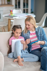 Obraz premium Dark-haired girl sitting on sofa, writing in her notebook, blonde female helping her, explaining something