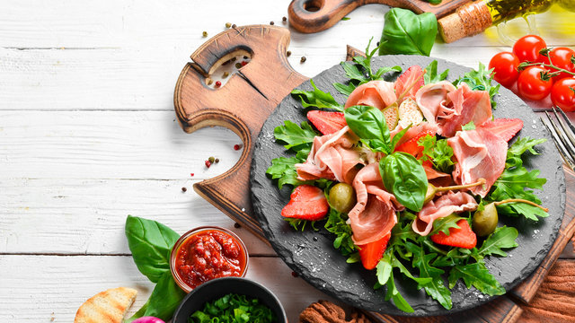 Salad of arugula, prosciutto, strawberries and capers on a black stone plate. Italian cuisine.
