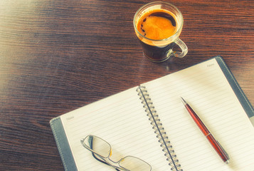 Cup of coffee, notebook with empty pages, pen, glasses in a metal frame on a table of brown wood.