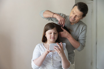 Caucasian dad learns to comb and trim daughter’s hair online. Father cuts the hair of a child to a teenager choosing a haircut in a smart phone, forgetful emotions and paternity, at home