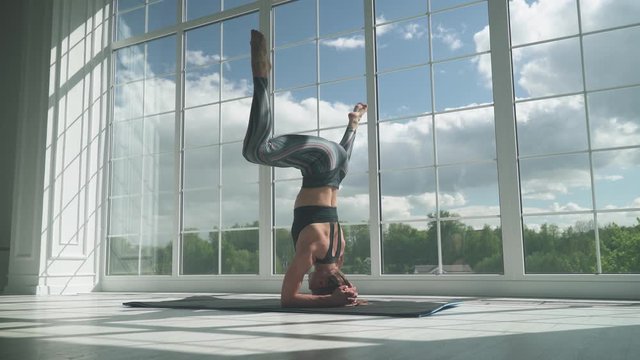 Young woman doing yoga in a white room filled with light, the girl performs a headstand near the large window.
