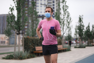 Sportsman with medical mask and gloves, smartphone and earbuds working out, jogging in urban surroundings.