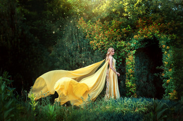 beautiful Russian folk princess is standing in fantasy forest. magic arch natural green leaves tree. Long blond braid hair. Woman queen. yellow elegant dress, cape cloak fly in wind. Сosplay rapunzel