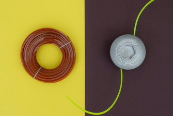 New metal reel for a lawn mower with a plastic yellow line on a dark background and new rolled brown line for a lawn mower on a yellow surface, top view.