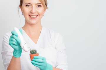 Cosmetologist is shaving green cactus in a brown pot with electric epilator wearing gloves smiling on white background. Concept of depilation, epilation and removal hair.