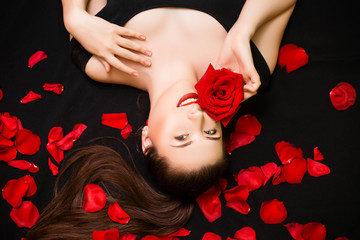 Portrait of a young, attractive woman with a rose in her hands lying on rose petals on a black background. Valentine's day concept. Model with retouched skin.