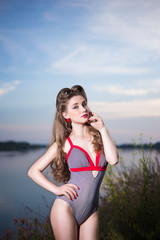 Pin up style portrait of a girl in striped swimsuit with long curly hair and red lipstick on river bank at summer day. Blue sky background, copy space.