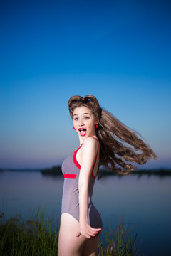Pin up style portrait of a girl in striped swimsuit with long curly hair and red lipstick, surprise emotion and flying hair on river bank at summer day. Blue sky background, copy space.