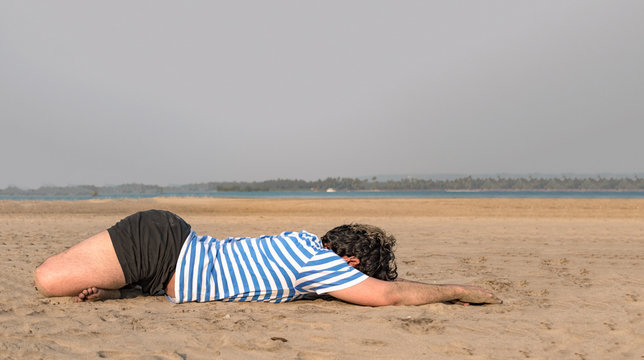 Side view photo of Young guy with fit body built practicing Yoga on beach in morning hours, by doing Forward bend Lotus position to keep his body flexible, relaxed and free from bone ailments. - Image