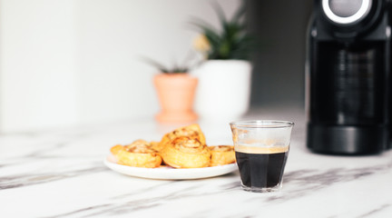 Close up picture of a fresh espresso in a glass cup next to a coffee machine and fresh baking buns. Marble table, cosy clean kitchen 