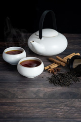 view of two cup of tea, teapot, cinnamon stick, tea strands, strainer, star anise on wooden table.