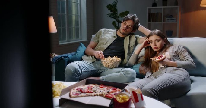 Caucasian extremely frustrated couple sitting in front of tv watching shows and eating pizza in absolute boredom. Student couple tired after a long day 4k footage