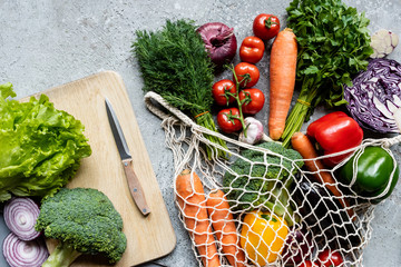 top view of fresh ripe vegetables in string bag near cutting board with knife on grey concrete surface
