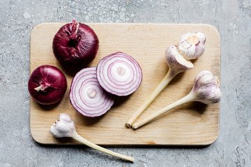 top view of onion and garlic on wooden cutting board on grey concrete surface