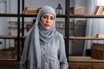 sad muslim woman in hijab looking at camera in living room, domestic violence concept