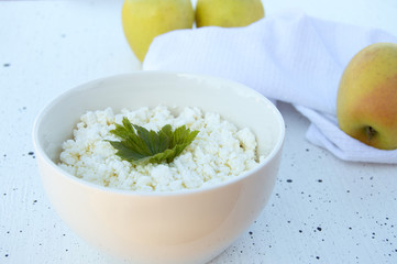 White bowl with homemade cottage cheese on a white background. Green apples in the background. Healthy diet.