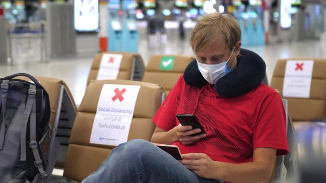 Traveling man wearing medical face mask at the airport uses phone and hold passport to prevent coronavirus infection during world epidemic