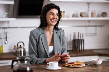 cheerful businesswoman using smartphone near breakfast and hiding problem of domestic violence