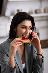 happy businesswoman eating toast bread with jam while talking on smartphone and hiding problem of domestic violence