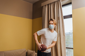 young man in medical mask holding american football in living room