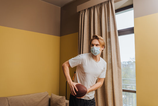 young man in medical mask holding american football in living room
