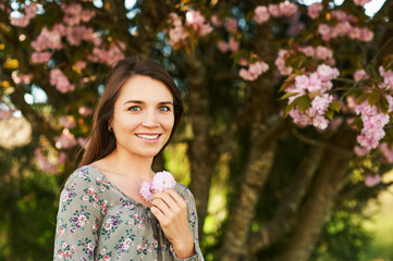 Outdoor spring portrait of pretty young woman with brown hair posing under pink blossoming tree