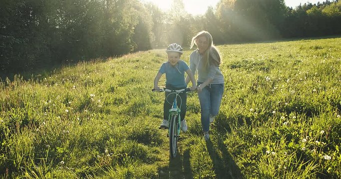 Mother teaching son to ride bicycle. Happy cute boy in helmet learn to riding a bike in park on green meadow in summer day at sunset time. Family weekend. 4K video Slow motion