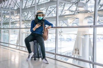 Asian female traveler wears a Corona-infected virus mask, sits on a suitcase, uses a smartphone to search for a route and check in International Airport.New normal ,Vacation and travel concept
