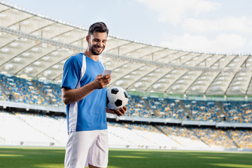 smiling professional soccer player in blue and white uniform with ball using smartphone at stadium © LIGHTFIELD STUDIOS