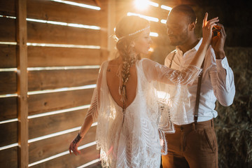 Newlyweds in the style of boho. African groom hugs a Caucasian bride in a barn in the sun. Flare effect. © MZaitsev