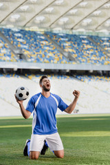 emotional professional soccer player in blue and white uniform with ball standing on knees on football pitch and showing yes gesture at stadium