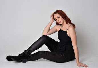 Fototapeta premium Portrait of a pretty girl with red hair wearing black leather pants, shirt and boots. full length sitting pose, isolated against a studio background