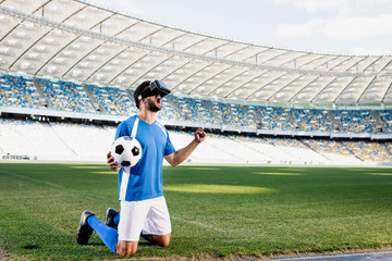 professional soccer player in vr headset and blue and white uniform with ball standing on knees and showing yes gesture on football pitch at stadium © LIGHTFIELD STUDIOS