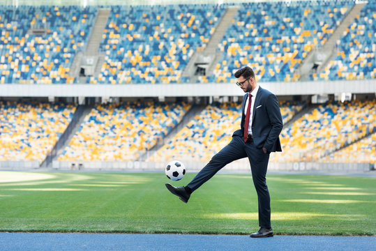 young businessman in suit and glasses playing with soccer ball at stadium