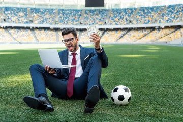 young businessman in suit with laptop, money and soccer ball sitting on football pitch at stadium...