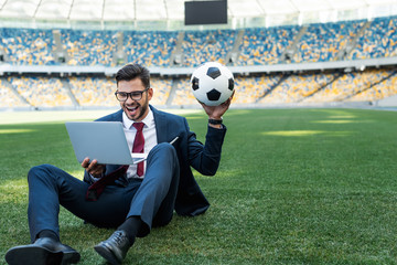 happy young businessman in suit with laptop and soccer ball sitting on football pitch at stadium, sports betting concept