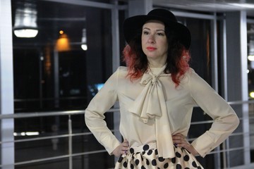 brunette in a beige blouse with a shirtfront and a skirt with black polka dots. a woman in a black hat with red hair against a glass wall with a railing, the night city outside the window. 