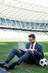 smiling young businessman in suit with laptop and soccer ball sitting on football pitch at stadium, sports betting concept