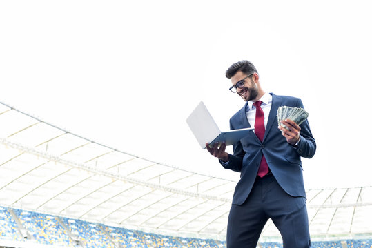 low angle view of happy young businessman in suit holding laptop and money at stadium, sports betting concept