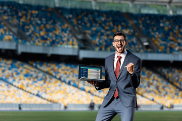 happy young businessman in suit holding laptop with sport betting website and showing yes gesture at stadium © LIGHTFIELD STUDIOS