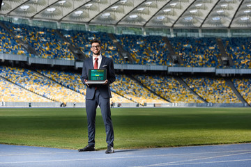 smiling young businessman in suit showing laptop with sports betting website at stadium