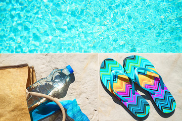 Swimming pool and straw hat, colorful flip flops, wicker bag, straw hat, bottle with still water as...