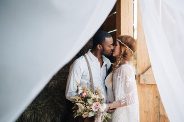 Newlyweds in the style of boho. African groom hugs a Caucasian bride in a barn.