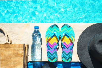 Swimming pool and straw hat, colorful flip flops, wicker bag, straw hat, bottle with still water as...